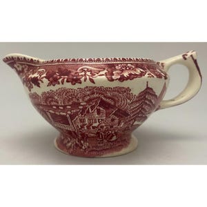 May include: A cream-colored ceramic creamer pitcher featuring a deep red transferware pattern. The design depicts a detailed rural landscape with houses, trees, and figures, complemented by a decorative floral border along the top rim and a handle.