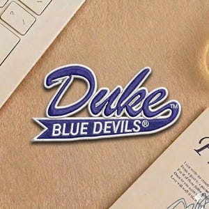 May include: A blue and white embroidered patch featuring the word "Duke" in a stylized script above the words "Blue Devils". The patch has a white border and is set against a tan background.