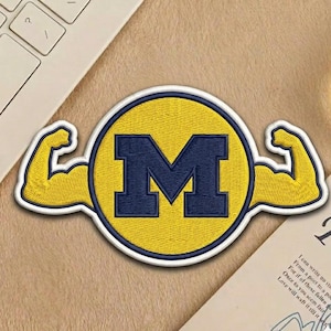 May include: Embroidered patch featuring a yellow circle with a navy blue "M" inside. Two yellow muscular arms flank the circle. The patch has a white border and is likely for apparel or accessories.
