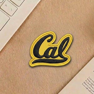 May include: A yellow and black embroidered patch with the word "Cal" in a script font. The patch has a yellow border and is likely for a college or university. The patch is on a tan surface.