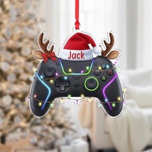 May include: A Christmas ornament shaped like a black video game controller, adorned with a Santa hat and reindeer antlers. The name "Jack" is printed on the hat. The controller is decorated with colorful lights and a red ribbon for hanging.