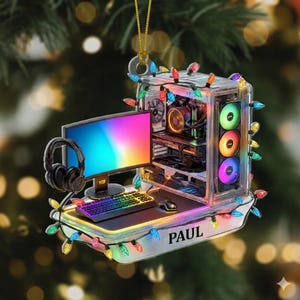 May include: A clear acrylic Christmas ornament shaped like a gaming PC setup. It features a monitor, keyboard, mouse, and headphones. The PC case has colorful fans and is decorated with string lights. The name "PAUL" is printed on the base.