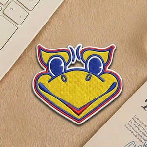 May include: Embroidered patch featuring a stylized yellow bird face with blue eyes and red and blue accents. The patch has a white border and is likely for application on clothing or accessories. The design is a cartoon-like representation.