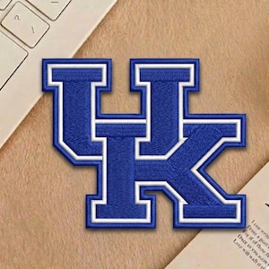 May include: Embroidered patch featuring the letters "UK" in blue with a white border. The patch is a stylized design, likely for apparel or accessories. The patch is on a tan surface, with a portion of a keyboard and a piece of paper visible.