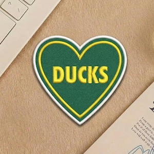 May include: A heart-shaped patch with the word "DUCKS" embroidered in yellow on a green background. The patch has a white border and a yellow outline. The patch is on a tan surface.