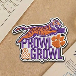 May include: Embroidered patch featuring a leaping tiger in orange and purple with the words "PROWL & GROWL". The patch includes an orange paw print and is outlined in white.