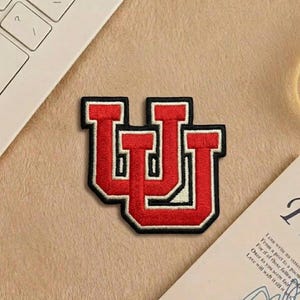 May include: Embroidered patch featuring a red "UU" design with a black border. The patch is on a beige surface, near a laptop keyboard and a card with text.