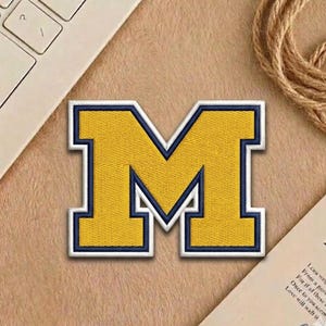 May include: Embroidered patch featuring a large, yellow letter "M" with a navy blue outline and accents. The patch has a white border and is likely for apparel or accessories. The patch is on a tan surface with a laptop keyboard and a piece of paper in the background.