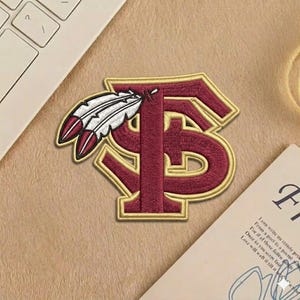 May include: Embroidered patch featuring the letters "FS" in maroon with a gold border, and a feather design. The patch is likely for apparel or accessories. The patch is on a tan surface.