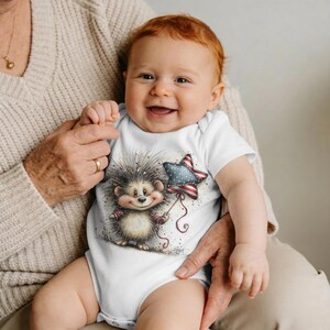 May include: A baby in a white onesie smiles, the garment featuring a cartoon hedgehog holding a star-shaped balloon. The balloon has a red, white, and blue pattern. The baby has red hair and is held by someone wearing a beige sweater.