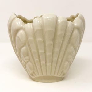 May include: A cream-colored ceramic planter shaped like a large seashell. The vessel features a glossy finish with vertical, raised ridges that mimic the natural texture of a scallop shell, creating a scalloped rim along the top edge of the bowl.