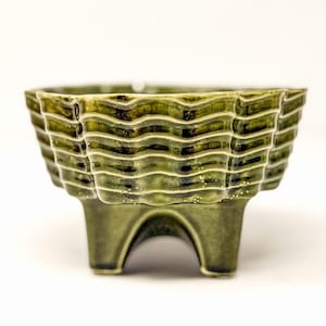 May include: A deep olive green ceramic bowl featuring a glossy finish and a textured exterior with horizontal wavy ridges. The bowl is supported by three short, sturdy feet that create arched openings between them at the base.
