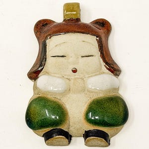 May include: A ceramic figurine of a seated figure with closed eyes, wearing a brown hat and robe. The figure has white accents on the chest and green accents on the lower body, with black shoes. The figurine has a small, rectangular handle at the top.