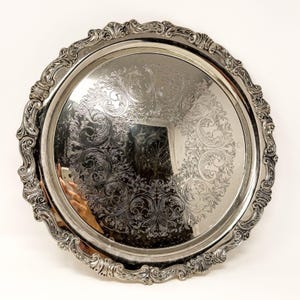 Peut inclure: Un plateau de service rond de couleur argentée doté d'un bord surélevé orné de volutes complexes et de motifs en forme de coquillages. Le centre du plateau présente une surface polie et réfléchissante décorée d'un motif floral gravé.
