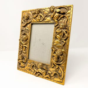 May include: A square gold-colored picture frame featuring a relief design of multiple cherubs playing various musical instruments amidst clouds. The frame has a dotted border pattern and a plain gray rectangular insert in the center.