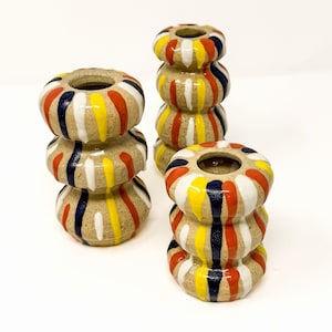May include: Three ceramic candle holders with a speckled beige base. Each holder features a stacked, rounded design decorated with vertical stripes in alternating colors of bright yellow, deep navy blue, vibrant orange, and crisp white paint.