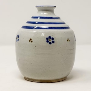 May include: A small, off-white ceramic vase with blue horizontal stripes around the neck and blue floral and brown dot designs. The vase has a rounded body and a narrow neck with a blue rim.