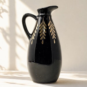 May include: A glossy black ceramic pitcher featuring a curved handle and a flared spout. The exterior surface is decorated with three vertical gold leaf-style patterns that extend downward from the neck of the vessel toward the rounded base.