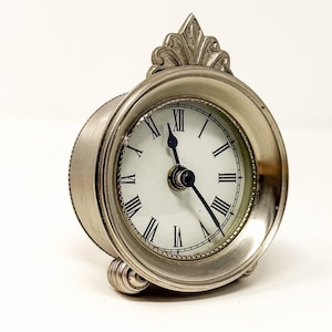 May include: A silver-toned metal table clock with a circular face, white dial, and black Roman numerals. The clock features a decorative finial at the top, two small rounded feet at the base, and two black hands indicating the time.