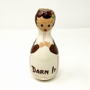 Könnte beinhalten: Eine Keramikfigur in Form einer Bowlingkegel mit bemaltem Gesicht, braunen Haaren und einer weißen Schürze. Die Figur hat braune Ärmel und den Schriftzug "Darn It" in braunen Buchstaben auf der Vorderseite der Schürze. Die Oberfläche ist glänzend.