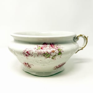 Op de afbeelding: Een witte keramische kamerpot met een bloemmotief van roze en groene bloesems. De pot heeft een brede, ronde vorm, een uitlopende rand en een enkel handvat aan de zijkant met goudkleurige accenten langs de randen van het handvat en de opening.