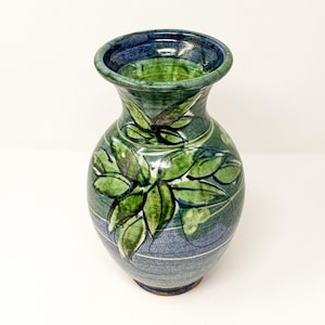 May include: A ceramic vase with a flared rim, featuring a hand-painted green leaf pattern over a blue and grey speckled glaze. The body of the vase has a rounded shape that tapers toward the base, with visible horizontal lines and brushwork.