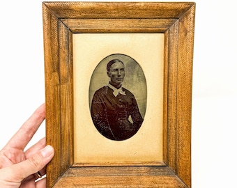 Può includere: Una cornice rettangolare in legno con una finitura testurizzata color marrone medio. La cornice contiene un passe-partout color crema con un'apertura ovale, che mostra un ritratto in bianco e nero di una persona con riga in mezzo e camicia.