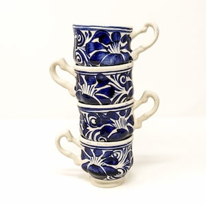 May include: A vertical stack of four ceramic cups, each featuring a white base with hand-painted deep blue floral and swirling patterns. Every cup has a distinctively curved, irregular handle and a slightly tapered shape, creating a uniform set.