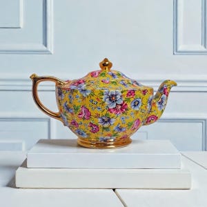 Könnte beinhalten: Eine gelbe Teekanne mit floralem Muster und goldenem Griff, Ausguss und Zierleisten. Die Teekanne hat einen Deckel mit einer goldenen Spitze und ist mit rosa, lila und blauen Blumen verziert. Die Teekanne steht auf einer weißen Oberfläche.