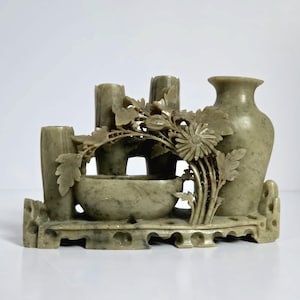 May include: A carved stone sculpture in shades of green and gray. The sculpture features a vase, several cylindrical shapes, and a small bowl, all connected by carved floral and leaf details. The piece is set on a decorative base.