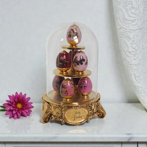 May include: A decorative display featuring six painted egg ornaments with floral designs, arranged on a tiered golden stand. The eggs are enclosed in a glass dome, and the base is adorned with ornate detailing and the text "Ruby Garden".