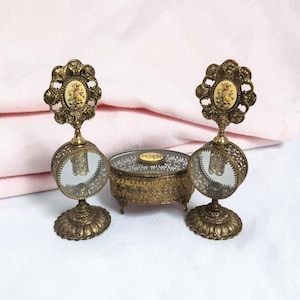 Puede incluir: Dos frascos de perfume de latón a juego con centros de vidrio circulares y tapones adornados con flores, acompañados de un pequeño joyero redondo de latón a juego con tapa de vidrio e incrustación floral central, todo con metalurgia detallada.