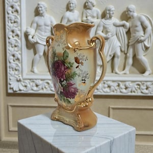 Könnte beinhalten: Eine antike Vase mit pfirsichfarbenem Sockel und Rand, verziert mit goldenen Akzenten und Griffen. Die Vase zeigt ein Blumendesign mit burgunderroten Rosen und grünen Blättern. Sie steht auf einem weißen Marmorwürfel, mit einem klassischen Relief im Hintergrund.