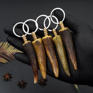 May include: Five antler keychains with brass accents and silver key rings. The keychains vary in color from dark brown to light yellow and green. The keychains are held in a gloved hand against a dark background.