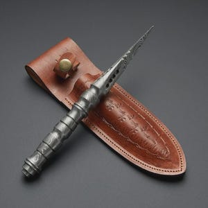May include: A handcrafted, ornate dagger with a patterned steel blade and handle, resting in a brown leather sheath. The sheath features decorative embossed detailing and a secure strap with a brass button. The dagger is a striking example of artisan craftsmanship.