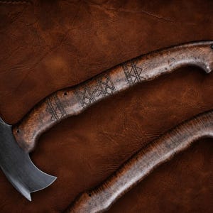 May include: A curved metal blade attached to a dark brown wooden handle featuring intricate geometric engravings. The handle has a smooth, polished finish with a small hole at the end. A second matching wooden handle is visible below the main tool.