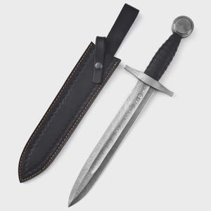 Puede incluir: Una daga de estilo medieval con una hoja estampada y un mango negro, junto a su funda de cuero negro. La daga presenta una guarda y un pomo redondeado. La funda tiene costuras decorativas.