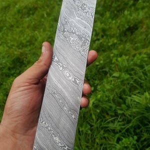 May include: A rectangular metal billet featuring a distinct gray and silver Damascus steel pattern with swirling, layered lines across the entire surface. The flat, elongated piece of metal displays a high-contrast, intricate grain structure.