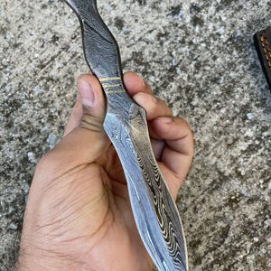 May include: A Damascus steel dagger with a patterned blade and handle. The handle has a curved design with gold-colored accents. The blade is sharp and pointed, and the overall color scheme is silver and black.