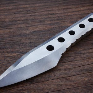 以下が含まれることがあります： 鋭く尖った刃と、上端に6つの円形の穴が並んだ銀色のナイフ。 ナイフは研磨仕上げで、濃い木目の背景に置かれています。 このデザインは、特定の目的のために作られたことを示唆しています。