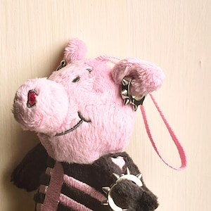 May include: A pink plush pig wearing a black outfit with a pink skeleton design. The pig has silver spiked earrings and a matching bracelet. A pink ribbon is attached to the pig's head.