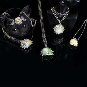 May include: A collection of silver-toned necklaces with pendant designs. The pendants feature light green and white shell-like stones, some with spiky silver metalwork. One necklace is displayed on a black cat-shaped holder, and others on a black mannequin.