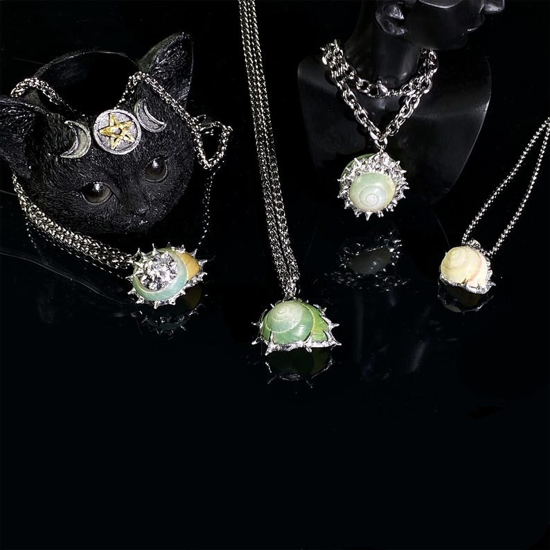 May include: A collection of silver-toned necklaces with pendant designs. The pendants feature light green and white shell-like stones, some with spiky silver metalwork. One necklace is displayed on a black cat-shaped holder, and others on a black mannequin.
