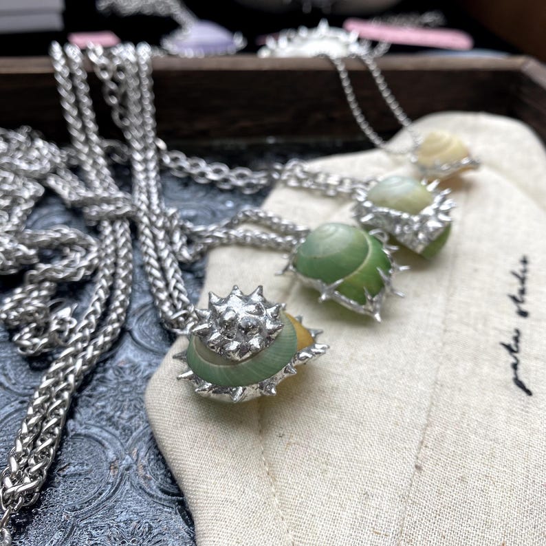 May include: Silver-toned necklaces with pendants shaped like sea shells. The shells are green and yellow, with a spiky silver metal border. The necklaces are displayed on a beige fabric.