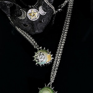 May include: A black cat sculpture with a silver and gold celestial headpiece. Two silver necklaces with pendant charms featuring a star and moon design. The pendants have a spiky, sun-like border and a moon-shaped accent.