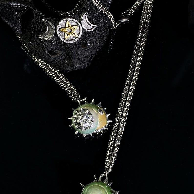 May include: A black cat sculpture with a silver and gold celestial headpiece. Two silver necklaces with pendant charms featuring a star and moon design. The pendants have a spiky, sun-like border and a moon-shaped accent.