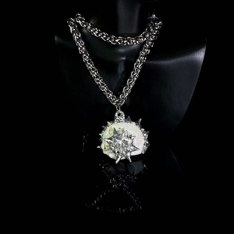 May include: A silver-toned necklace with a large pendant. The pendant is a white, oval-shaped stone with a spiky, starburst-like silver metal design. The chain is a thick, textured silver. The necklace is displayed on a black mannequin.