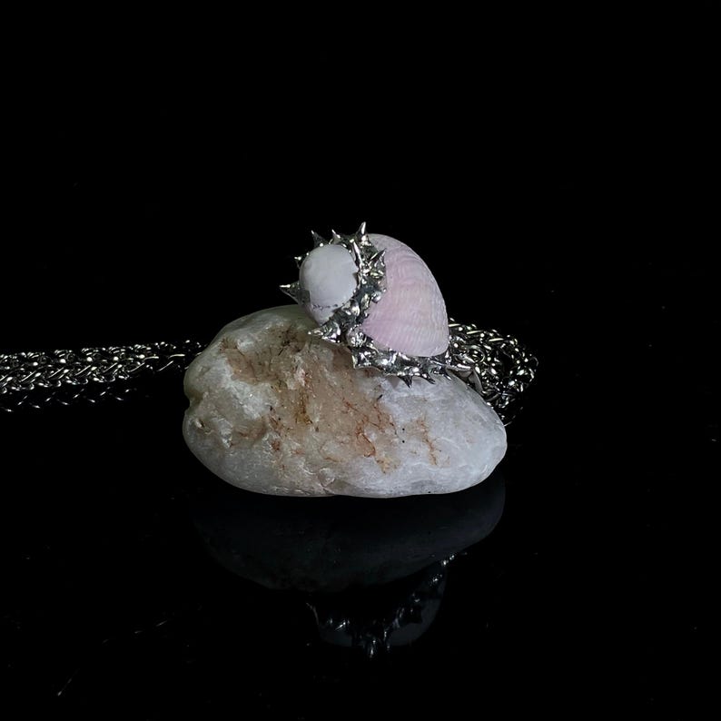 May include: A unique necklace featuring a light pink seashell adorned with a silver-toned, spiky ring. The seashell rests on a white stone, with a silver chain visible. The background is black, highlighting the jewelry.