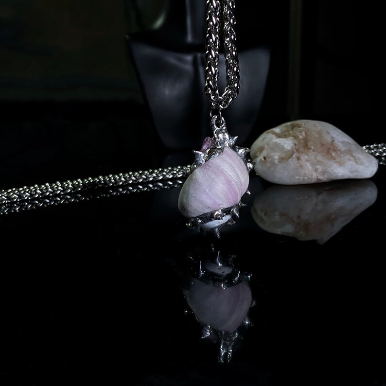 May include: A silver-toned necklace featuring a pendant made from a light pink, striped stone. The stone is encased in a silver metal setting with spiky accents and a small purple stone. The necklace is displayed on a reflective black surface.