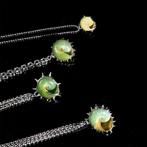 May include: Four necklaces with pendants shaped like spiral seashells. The shells are a mix of green and yellow, set in silver-colored metal. The chains are also silver-colored and have a ball chain design. The background is black.